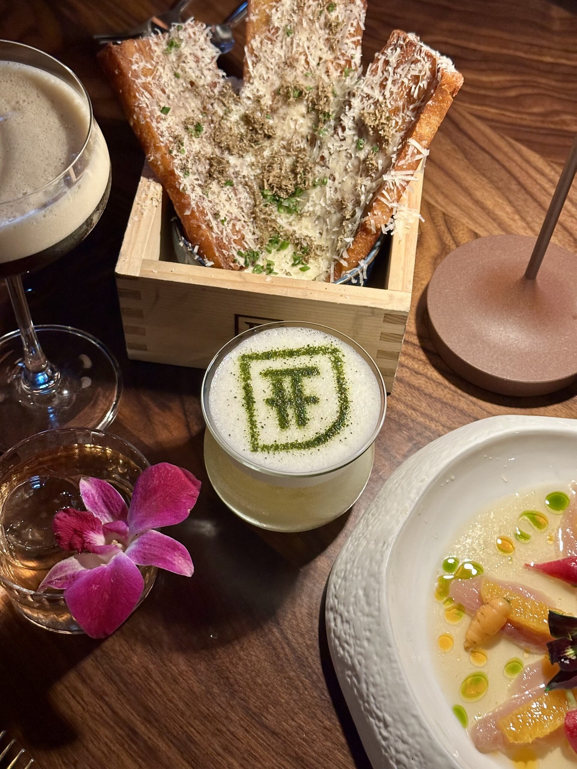 A matcha cocktail, crispy potato appetizer, flowery cocktail and raw hamachi appetizer are in frame on a wooden table.