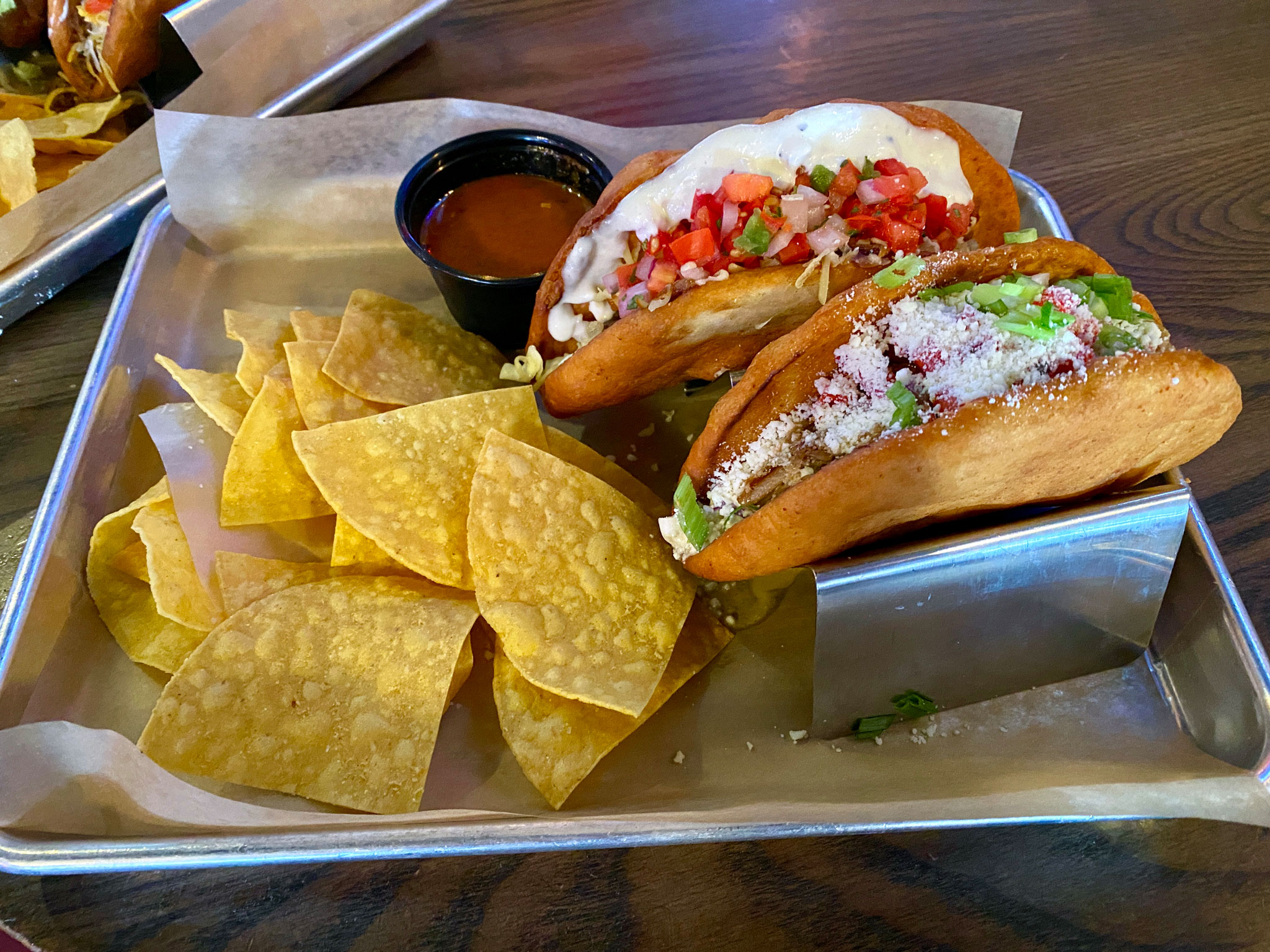 Chalupas - Smokey and The Brisket and Porkin' It Back: Smokey and The Brisket - Smoked brisket, BBQ sauce, smoked gouda, pico de gallo, poblano ranch slaw, smoked onion aioli, pickled red onions. Front: Porkin' It - Slow and low pork carnitas, Korean BBQ, pineapple salsa, cotija, pico de gallo, biggity bang aioli slaw, cilantro.